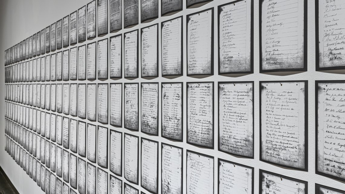 A sidelong view of Deanna Bowen's prints of a 1911 petition, showing black-edged letter-sized pages receding in rows along a white gallery wall