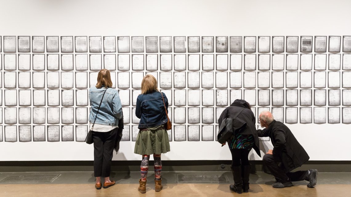Photo of two pairs of gallery visitors seen from behind as they examine rows of pages of Deanna Bowen's installation of a 1911 petition, which fills the wall to both edges of the photo