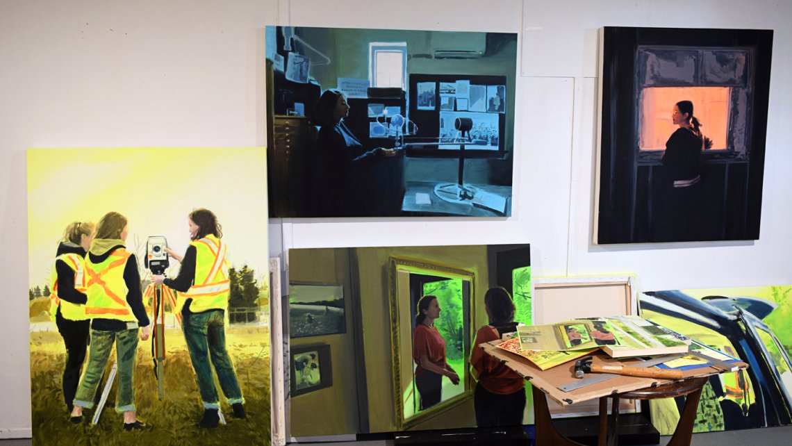 View of Amanda Rhodenizer's studio wall showing four paintings in progress of working women in saturated colour settings; a small studio table sits in the foreground