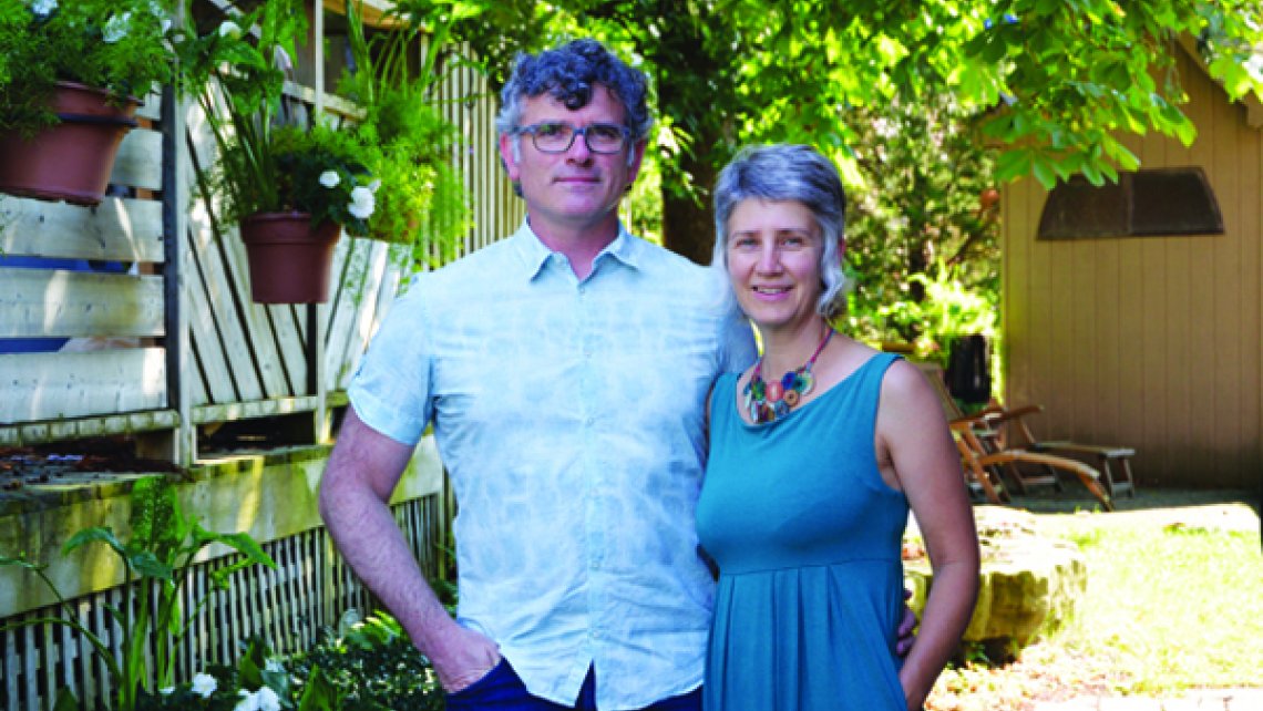 Photo of Trent Bauman and Juanita Metzger, an older couple casually dressed and standing together in a backyard filled with lush greenery