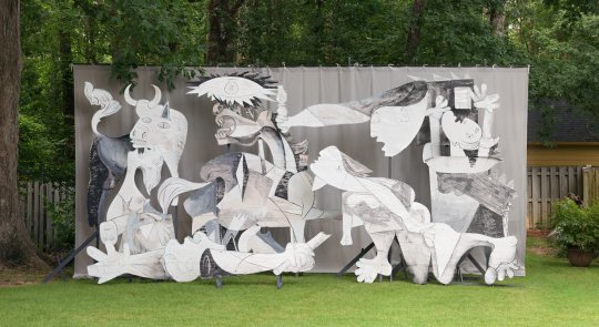Adad Hannah photo of a restaging of Picasso's Guernica using wood and cardboard cut-outs, arranged in a lush green backyard space