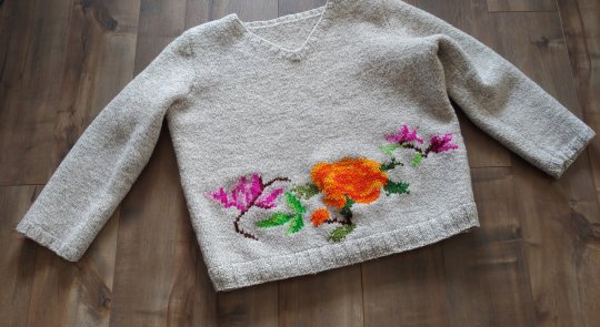 A white knitted sweater spread out on a dark wood laminate floor with a pattern of violet and orange flowers worked into its lower edge