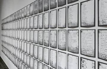 A sidelong view of Deanna Bowen's prints of a 1911 petition, showing black-edged letter-sized pages receding in rows along a white gallery wall